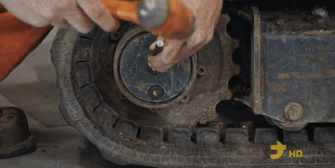 A heavy-duty mechanic uses a chisel and hammer to loosen a final drive drain plug