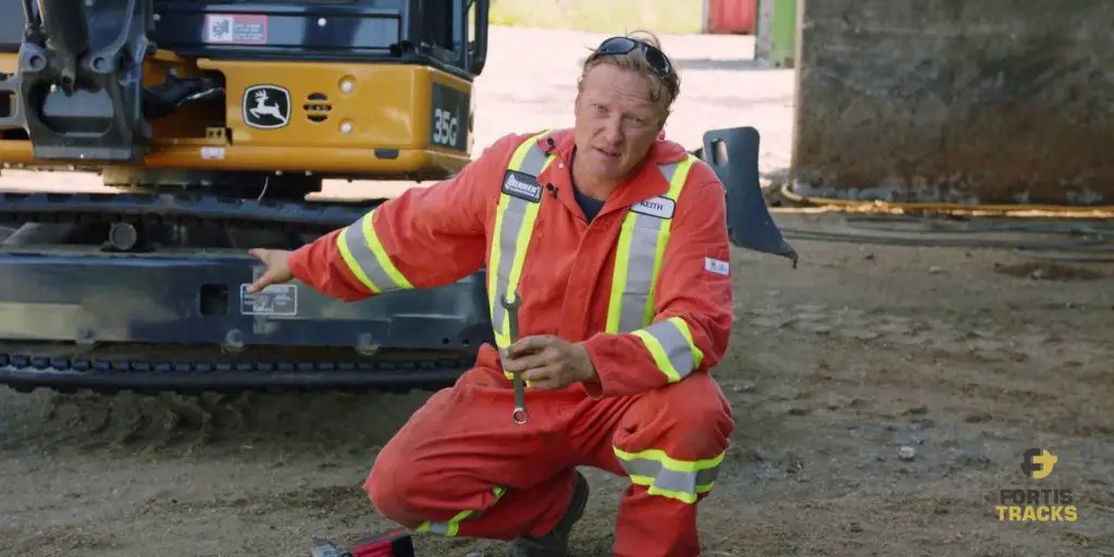 Heavy duty mechanic holding am open-ended wrench while pointing at a mini excavator behind him