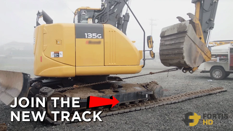 A John Deere 135G Excavator runs down on a new steel track while the bucket pulls on one of the track’s ends with a chain.