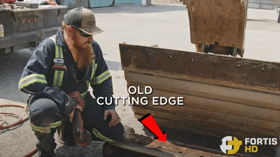 A heavy-duty mechanic is beside an excavator bucket with a detached cutting edge.