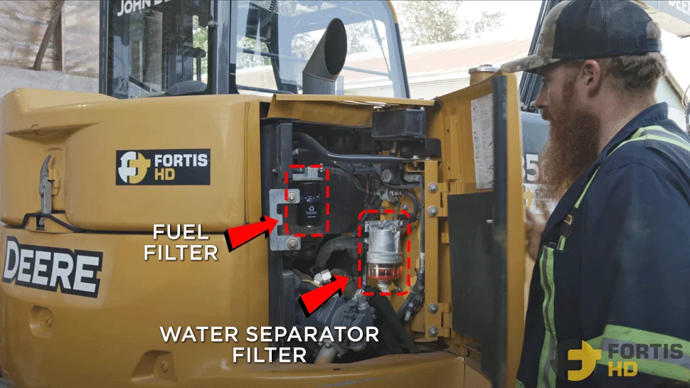 A heavy-duty mechanic opens the right compartment on a John Deere 85G to show the fuel and water separator filters.
