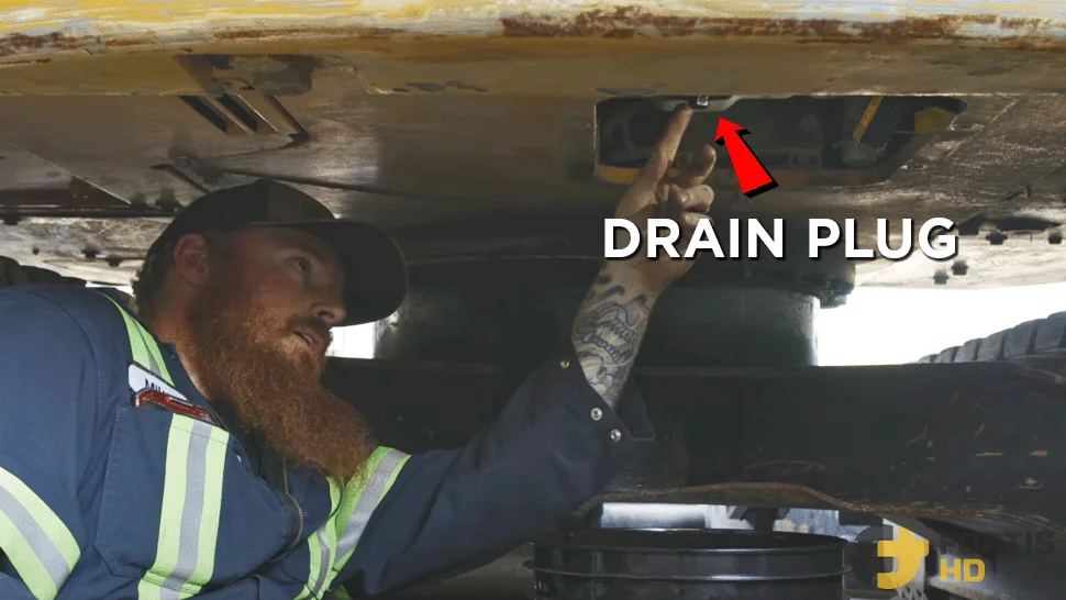 Heavy duty mechanic underneath a John Deer 85G Excavator. An overlay arrow shows the position of the drain plug.